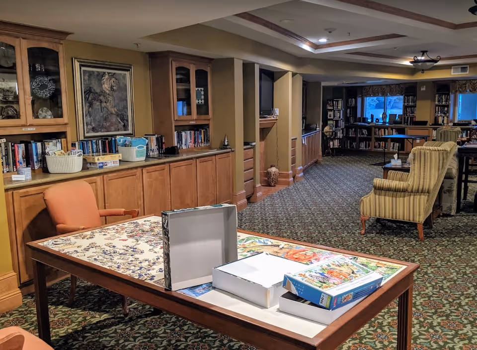 Carpeted community lounge with a table holding a partially assembled puzzle and boxes, bookshelves along the wall, and upholstered chairs.