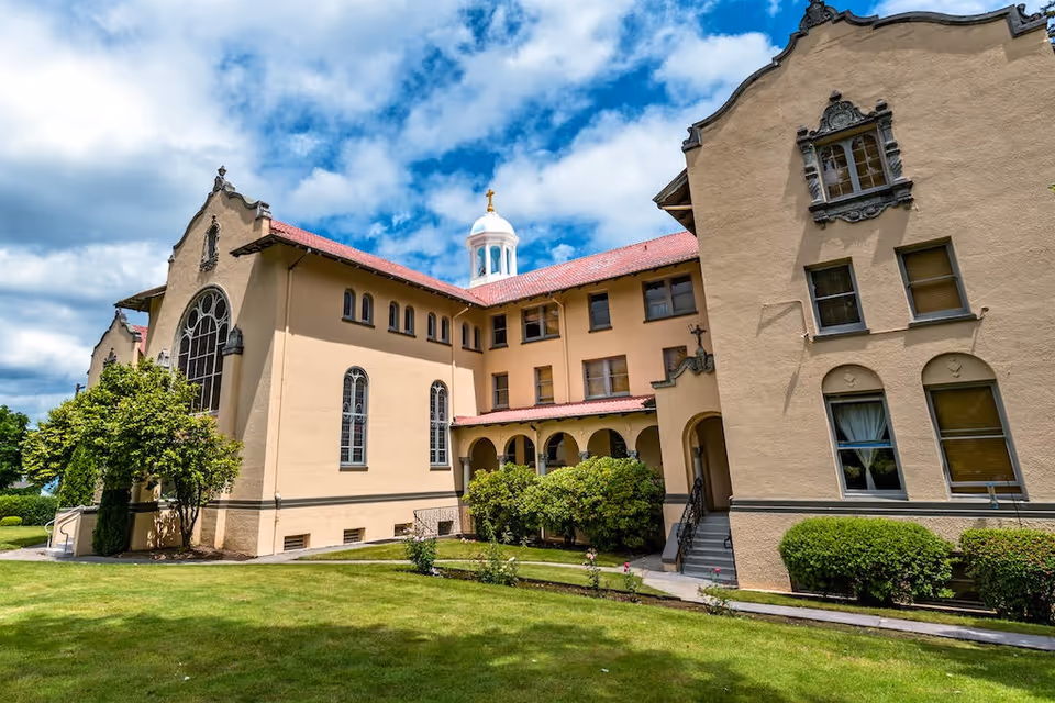 Exterior view of a large, beige stucco building with red tile roofing and arched windows, surrounded by green grass and bushes under a partly cloudy blue sky.