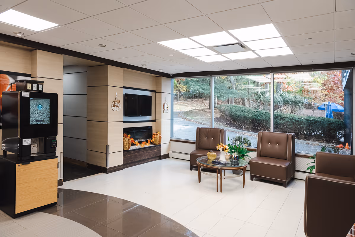 A bright and modern seating area with large windows showing an outdoor view of trees and bushes. The room features brown cushioned chairs arranged around a glass coffee table with decorative plants and flowers. On the left side, there is a coffee machine, and the back wall has a mounted flat-screen TV above a decorative fireplace with autumn-themed decorations.