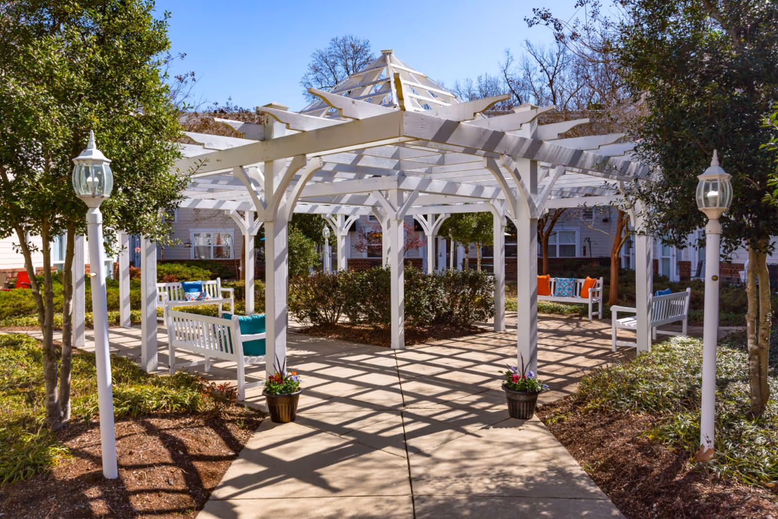 Outdoor seating area with white wooden pergola structure, white benches with colorful cushions, potted plants, and surrounding greenery under a clear blue sky.