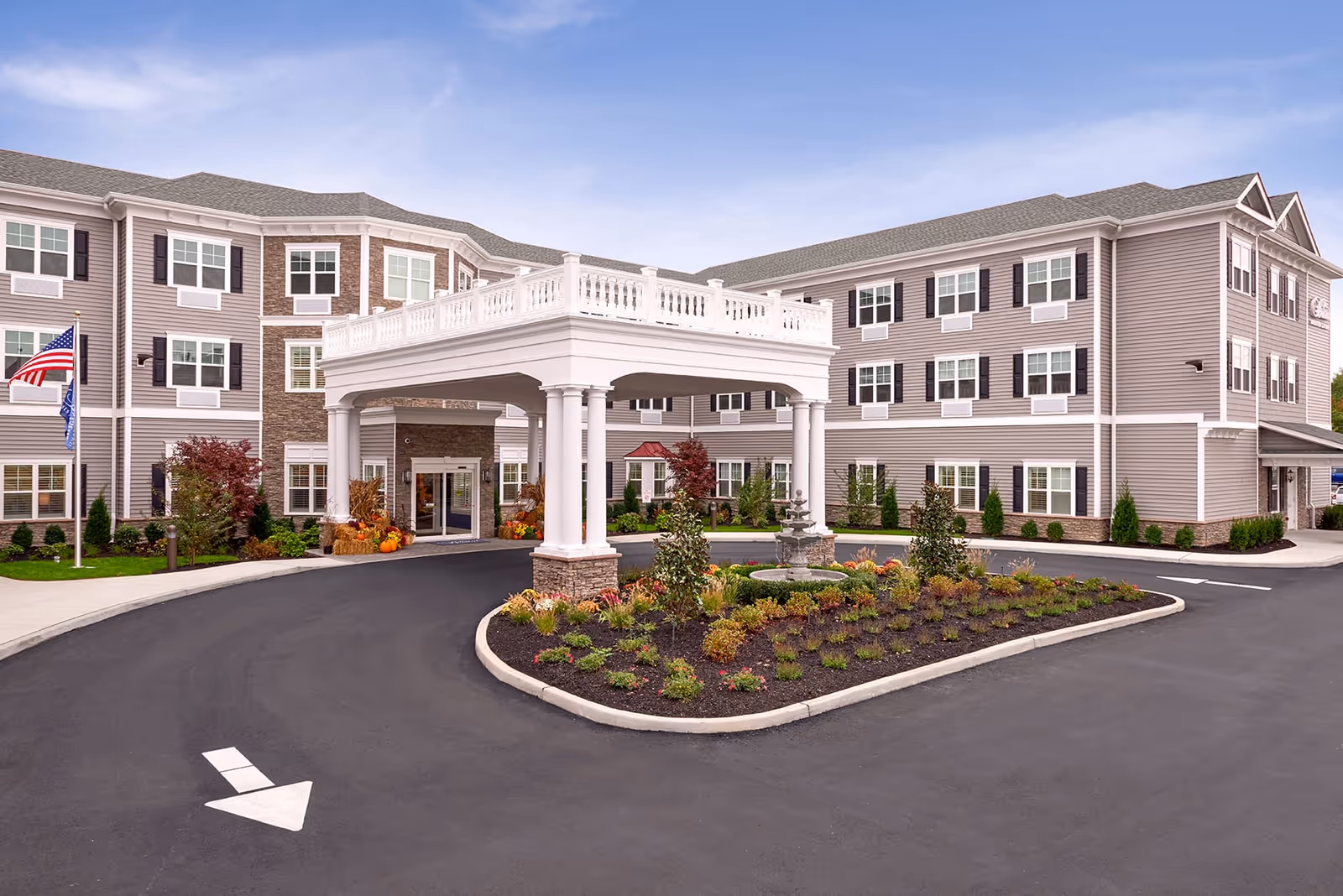 Exterior view of The Bristal Assisted Living at West Babylon building with a covered entrance, landscaped garden with a small fountain, and American flags near the entrance.