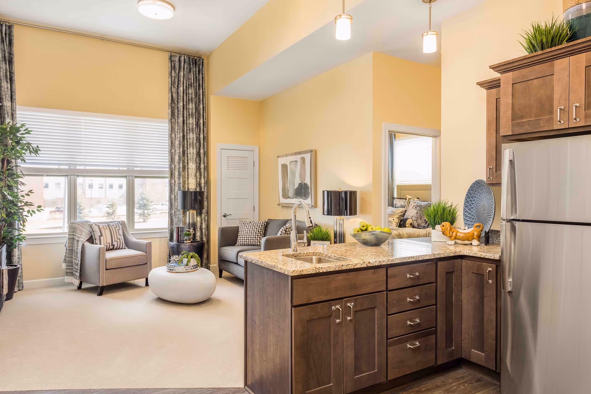 A bright and cozy living area with a kitchen island in the foreground featuring a granite countertop, a sink, and wooden cabinets. The living room has a gray armchair with a throw blanket, a small round white coffee table, a gray sofa with patterned pillows, and two black table lamps. Large windows with blinds and patterned curtains allow natural light to fill the space. A doorway leads to a bedroom with a bed visible. Decorative plants and artwork add a homey touch.