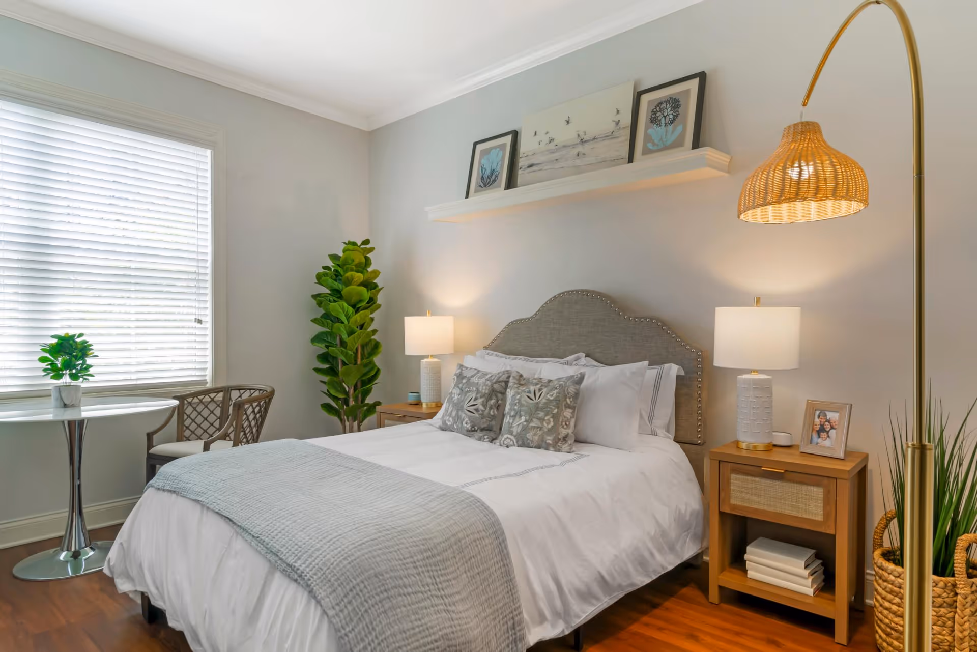 A cozy bedroom with a neatly made bed featuring white and gray bedding and decorative pillows. There are two wooden nightstands on either side of the bed, each with a lamp. A tall green plant is placed in the corner near a window with white blinds. A small round table with two chairs is positioned by the window. Above the bed, a white floating shelf holds three framed artworks. A woven floor lamp stands next to the nightstand on the right, and a basket with greenery is on the floor beside it.