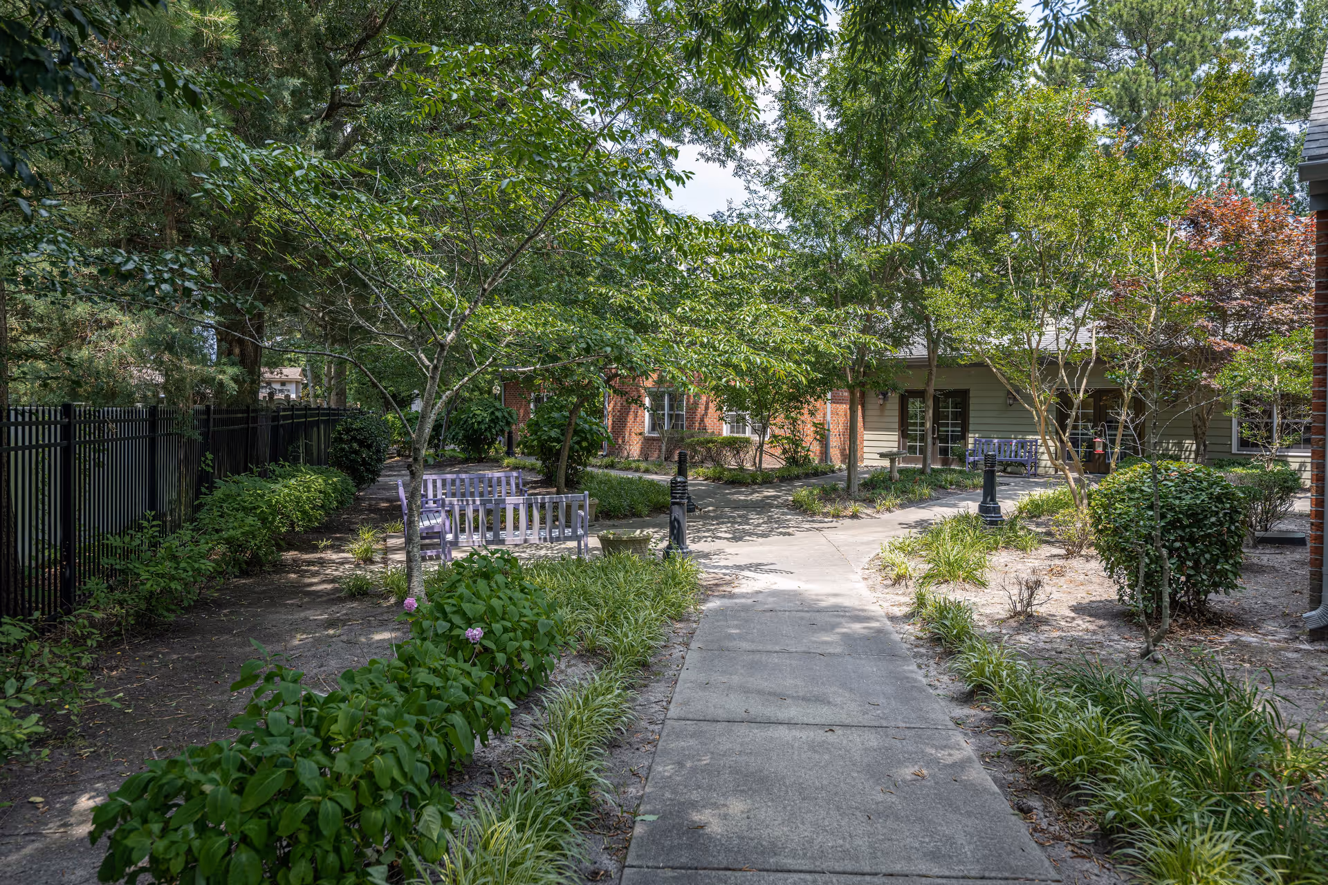 A peaceful outdoor garden area with a concrete pathway leading through green bushes and trees. There are several purple benches along the path and a black metal fence on the left side. The background shows parts of a building with brick and siding walls.