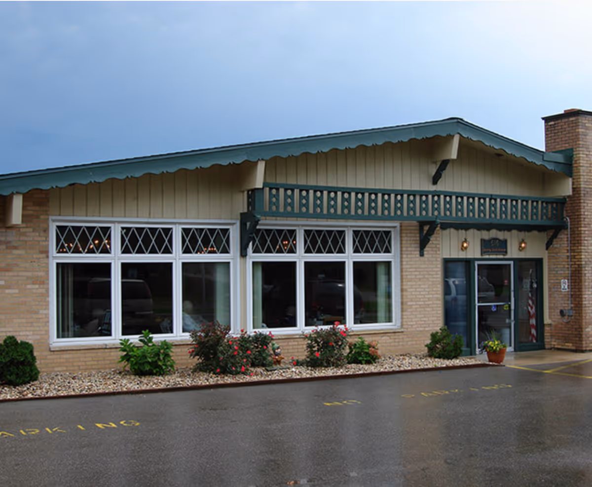 Front exterior of a single-story healthcare building with large diamond-pane windows, a decorative green awning, entrance door, and small landscaped beds by a wet driveway.