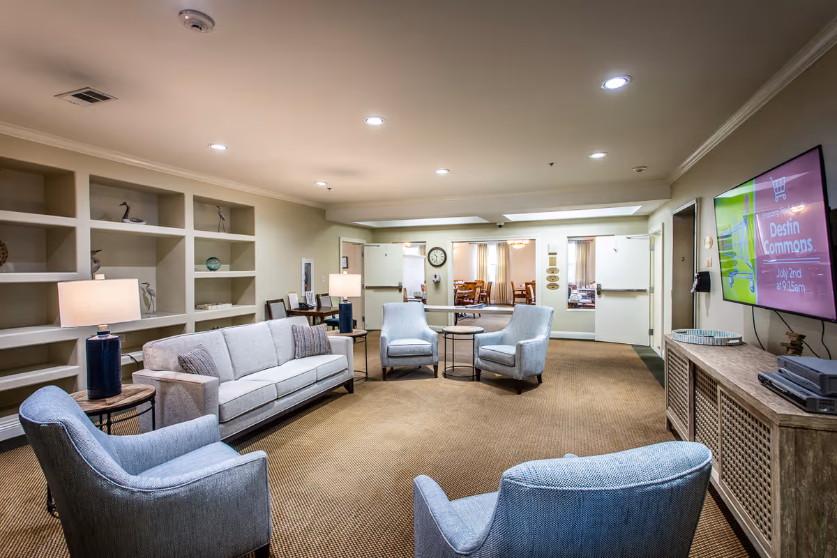 Bright communal living room with sofas and armchairs arranged around side tables, built-in shelving, lamps, and a wall-mounted TV.