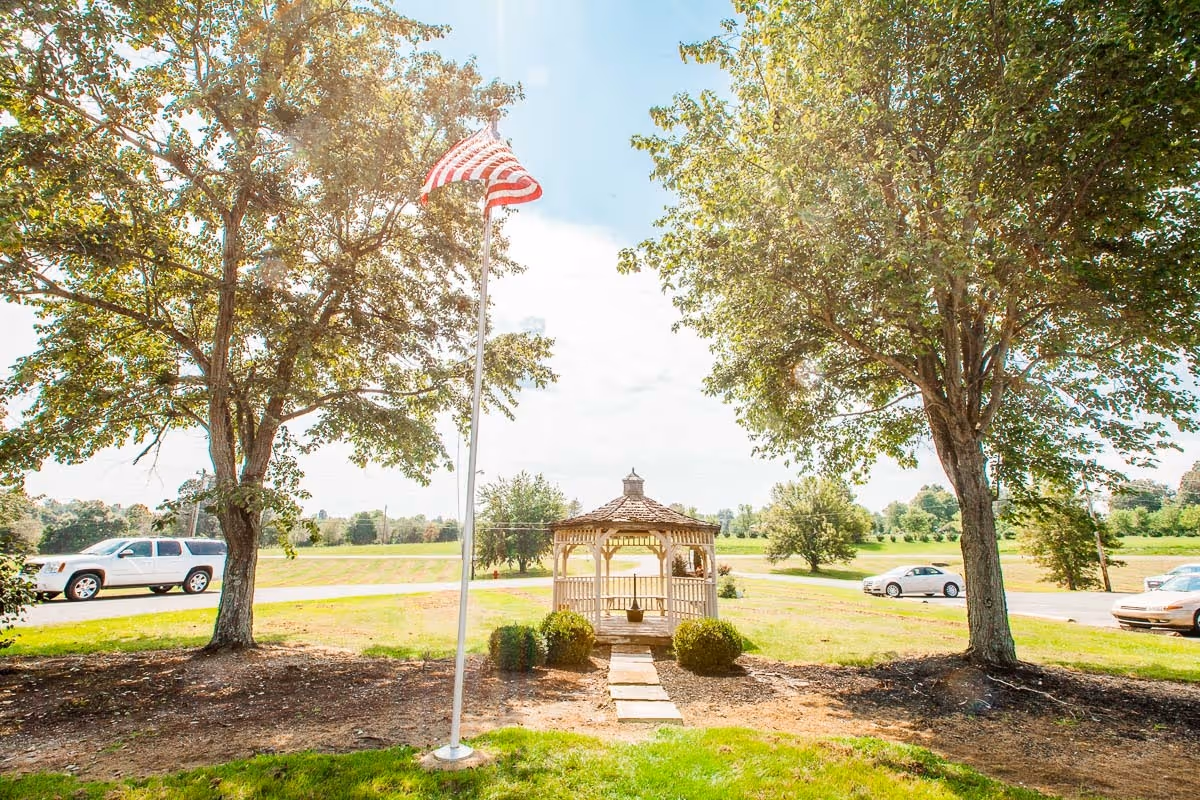 A landscaped outdoor lawn with a gazebo centered between two trees and an American flag on a pole.