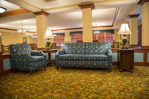 A cozy seating area in a senior living facility featuring a blue floral-patterned armchair and matching sofa. The furniture is placed on a patterned carpet with warm tones. Two wooden side tables with lamps flank the sofa. In the background, there are dining tables and chairs under hanging chandeliers, with large windows covered by blinds.