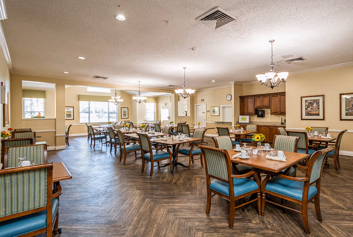 Spacious dining room with multiple set wooden tables and upholstered chairs, chandeliers, and a serving area in the background.