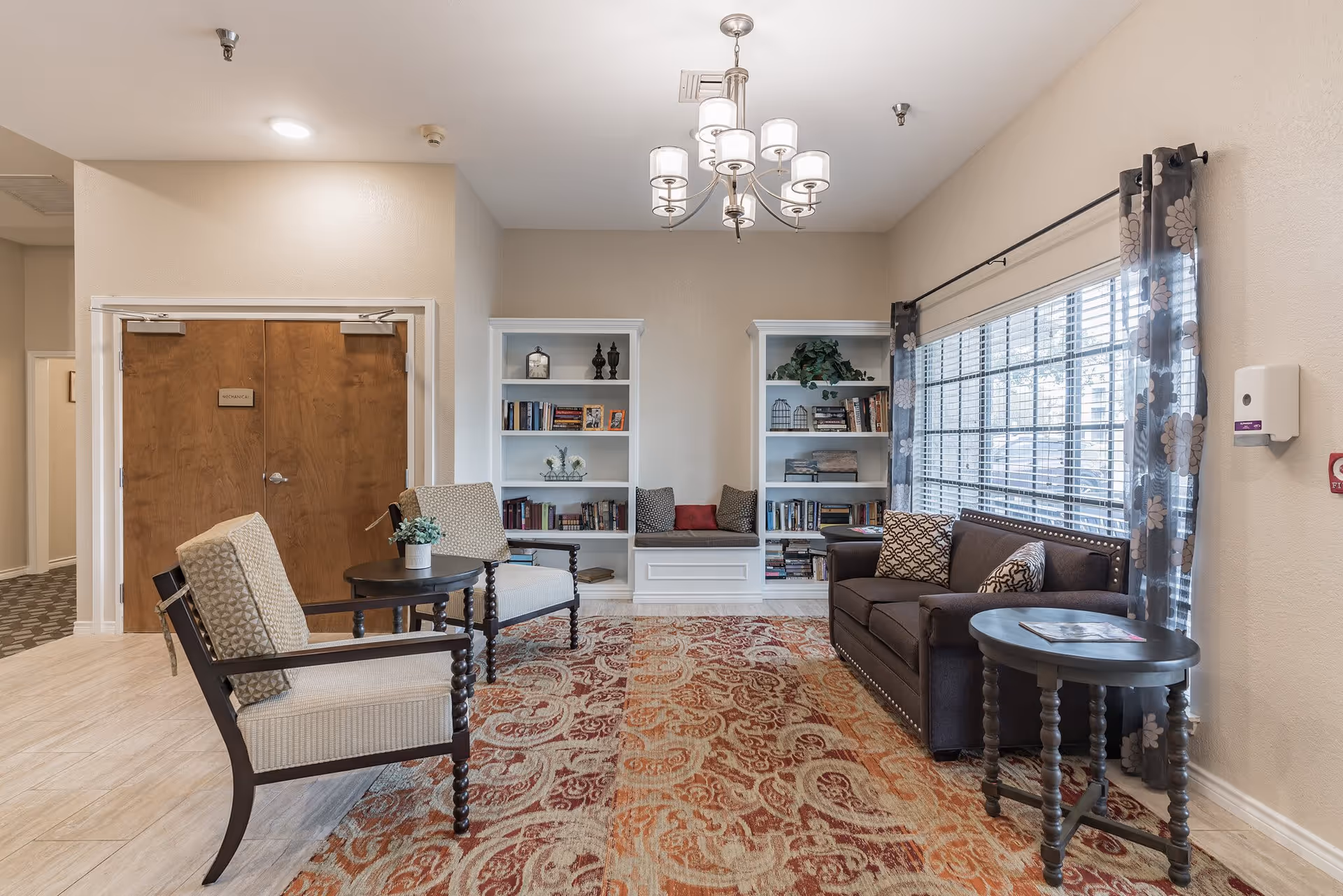 A cozy common room with armchairs, a sofa, built-in bookshelves, a patterned rug and a large window.