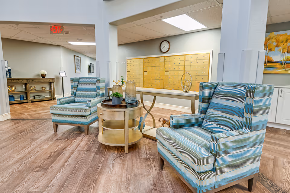 A senior living facility lounge with patterned armchairs, a round wooden side table, and a wall of mailboxes behind a console table.