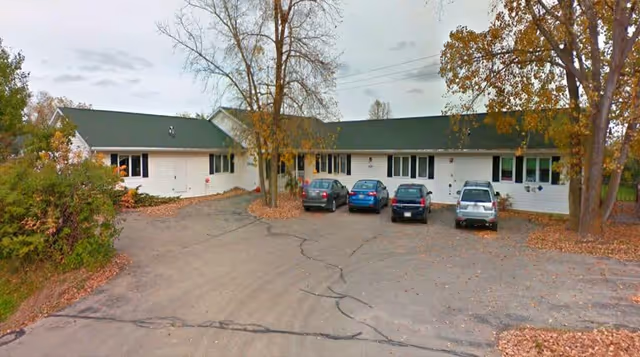 Single-story white building with a green roof surrounded by trees with autumn leaves. Four cars are parked in front on a cracked asphalt driveway. The scene appears to be an exterior view of a residential or care facility.