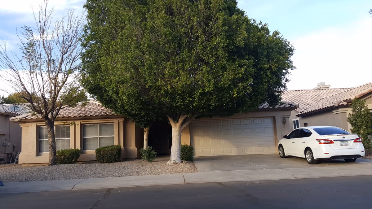 Single-story stucco house with a tiled roof, large front tree, two-car garage and a white car parked in the driveway.