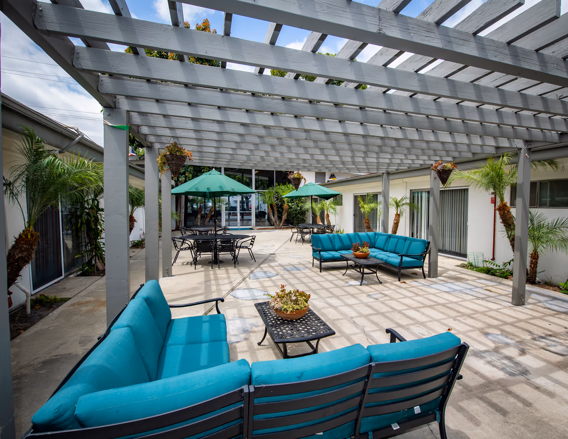 Shaded outdoor courtyard with teal patio sofas, tables, umbrellas, and a wooden pergola.