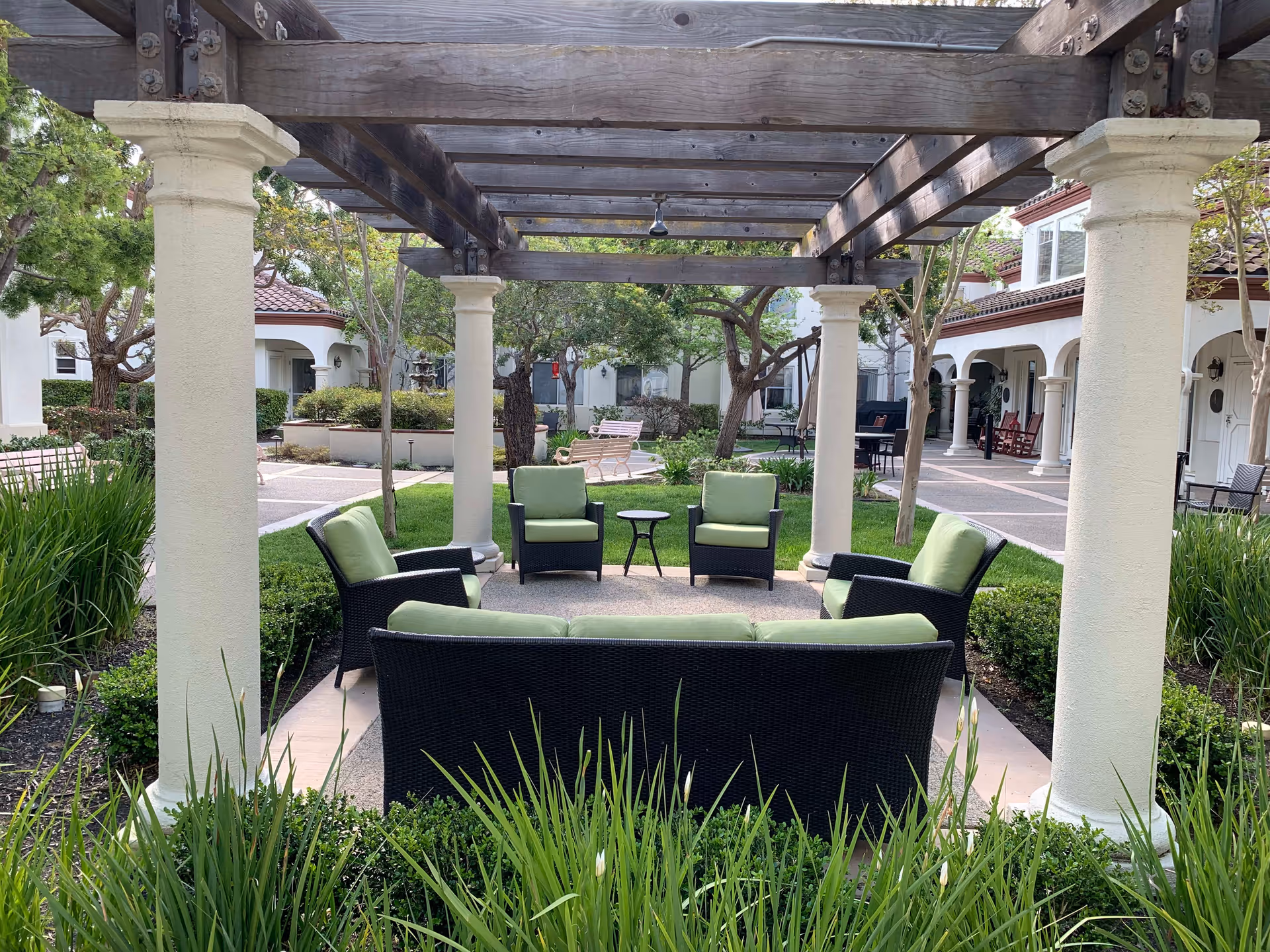 Outdoor seating area under a wooden pergola with four armchairs and a sofa with green cushions arranged around a small round table. The area is surrounded by greenery and bushes, with a paved walkway and white buildings with arches and red-tiled roofs in the background.