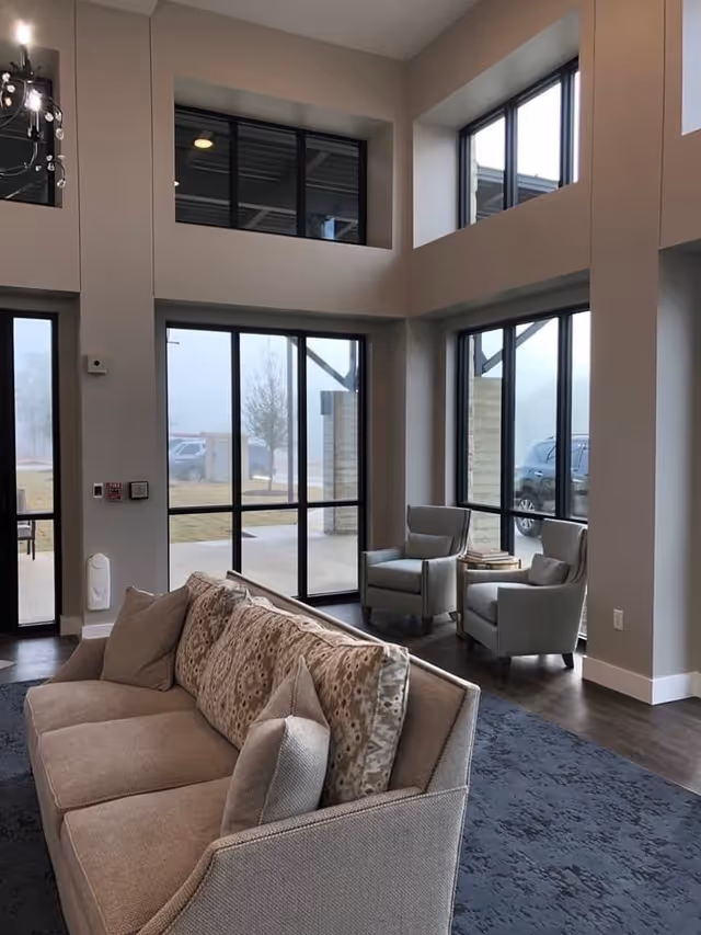 A modern living room area with a beige sofa adorned with patterned and plain cushions, two gray armchairs positioned near large floor-to-ceiling windows, and a small round table between the chairs. The room features high ceilings, dark wood flooring, and a blue patterned rug. Outside the windows, a parking area and some trees are visible on a foggy day.