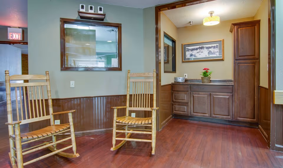 Interior view of a room with two wooden rocking chairs on a polished wooden floor. There is a window with a wooden frame on the wall behind the chairs, and a countertop with wooden cabinets on the right side. A framed picture and a potted plant with red flowers are on the countertop. An exit sign is visible in the background on the left.