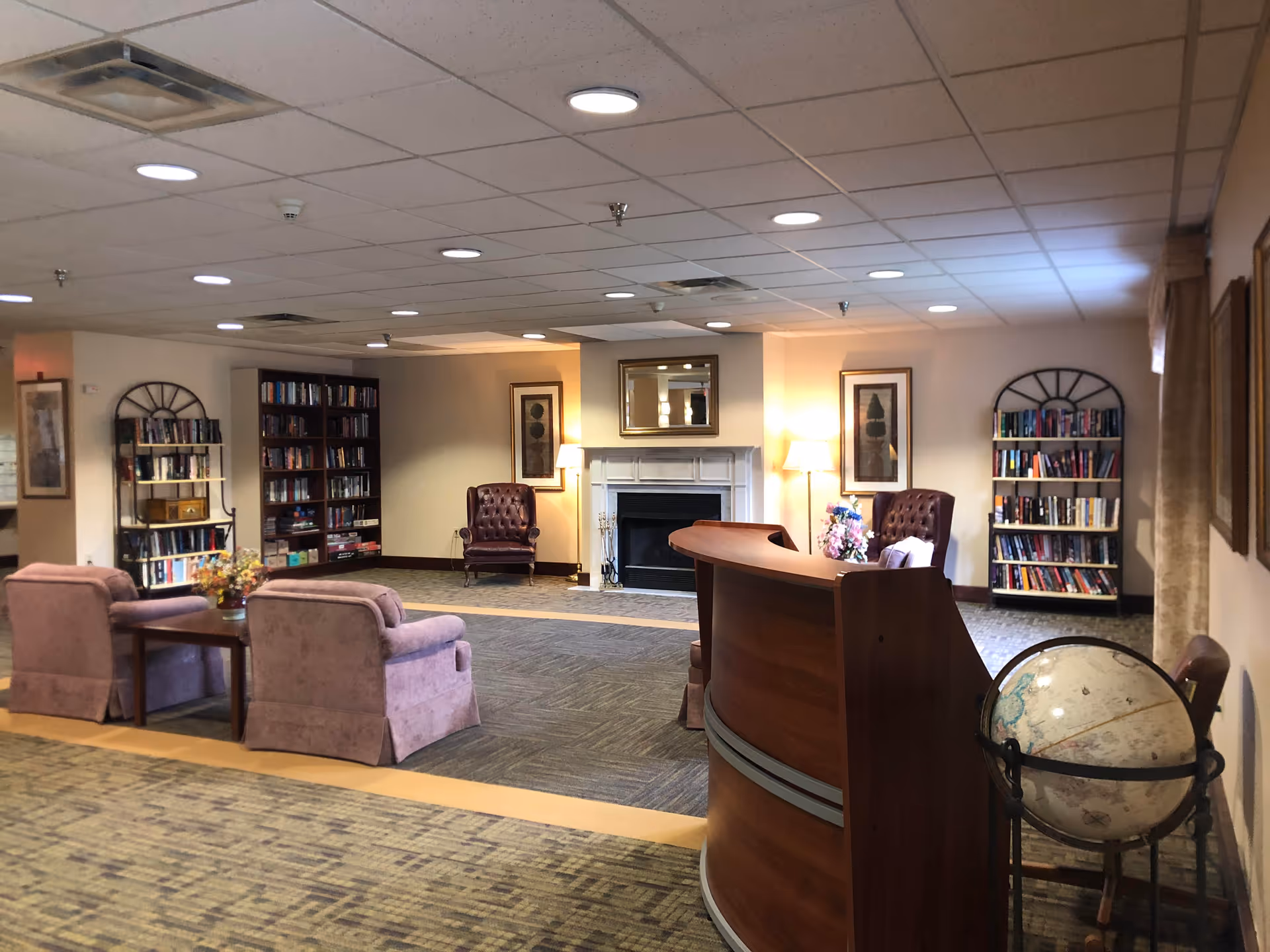 Carpeted common lounge with bookshelves, armchairs, a fireplace, and a curved reception desk.