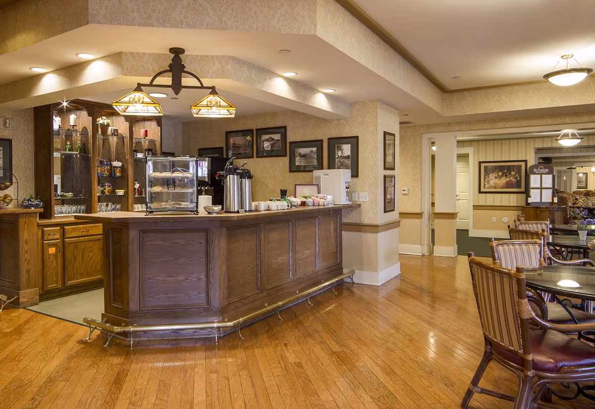 Interior view of a senior living facility's coffee and snack bar area with wooden cabinetry, a counter with coffee dispensers, cups, and a display case with pastries. To the right, there are tables and chairs with striped upholstery in a dining or common area. The room has warm lighting, hardwood floors, and framed pictures on the walls.