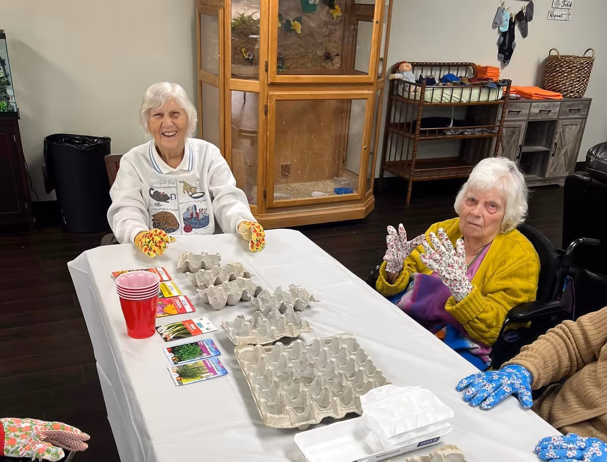 Two elderly women sitting at a table covered with a white tablecloth, wearing colorful gardening gloves. The table has several empty egg cartons and seed packets arranged on it. One woman is smiling and the other is looking at the camera. In the background, there is a wooden cabinet with glass doors, a changing table with folded towels, and a wicker basket.