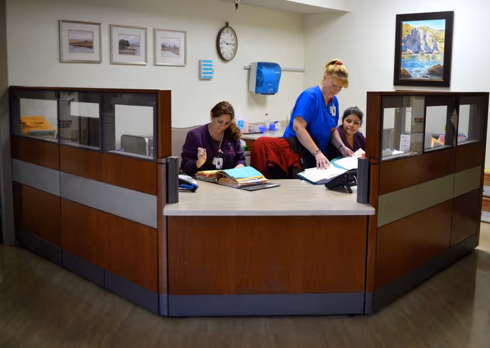 Three staff members working at a reception desk in an office area. Two are seated and reviewing documents, while one stands and points at a file. The desk is wooden with glass partitions, and the background has framed pictures, a clock, and a wall-mounted paper towel dispenser.