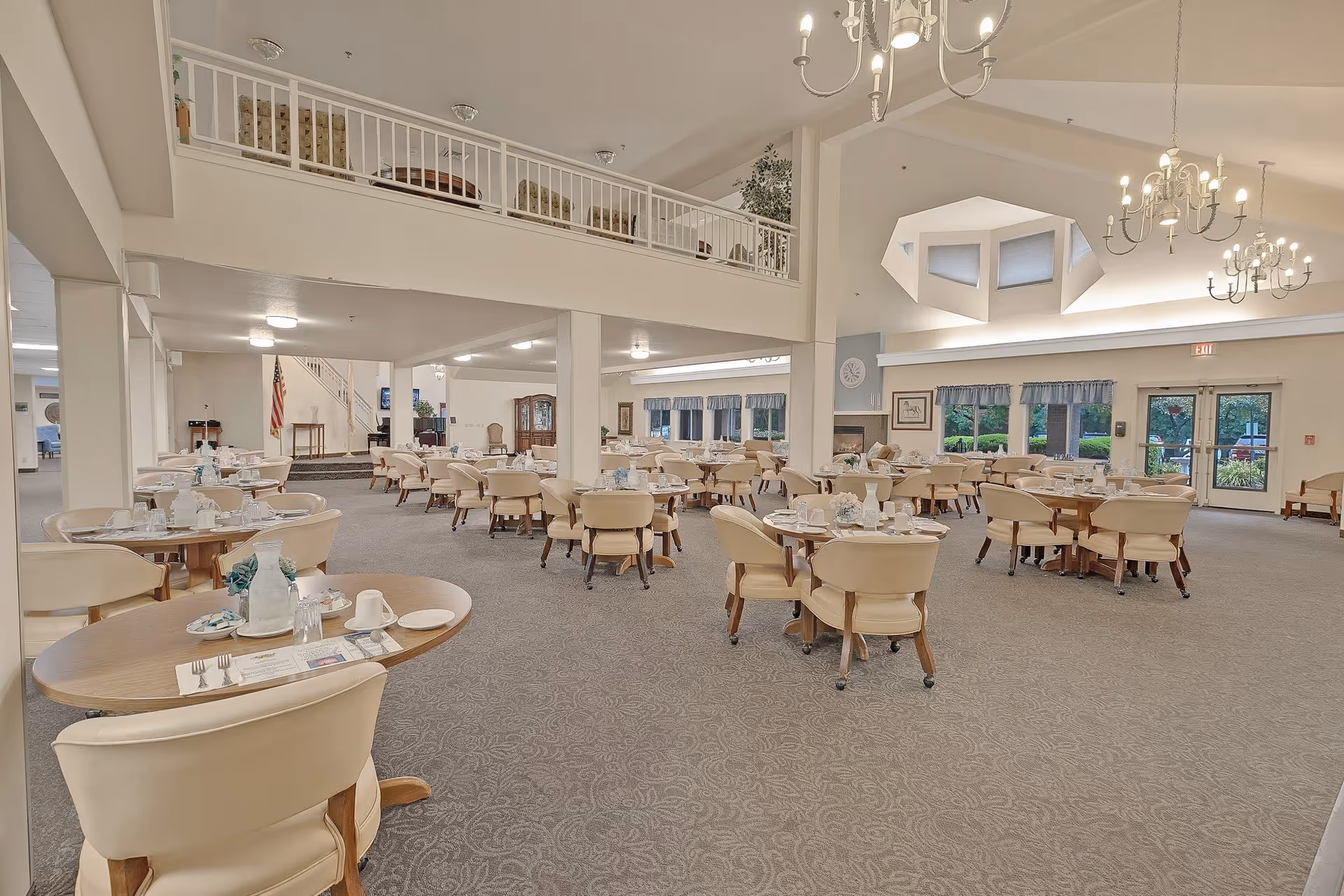 Spacious dining room in a senior living community with multiple round tables set with plates, cups, and utensils. The room features beige upholstered chairs, carpeted floors, large windows with blue valances, chandeliers hanging from a high ceiling, and a balcony area above.