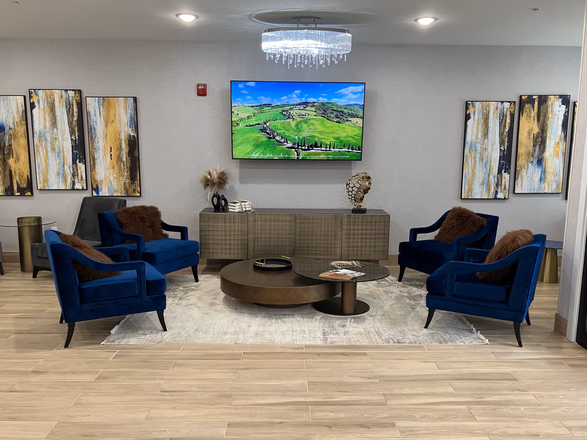 A modern common room with four blue velvet armchairs around round coffee tables, a wall-mounted TV, abstract artwork, and a chandelier.