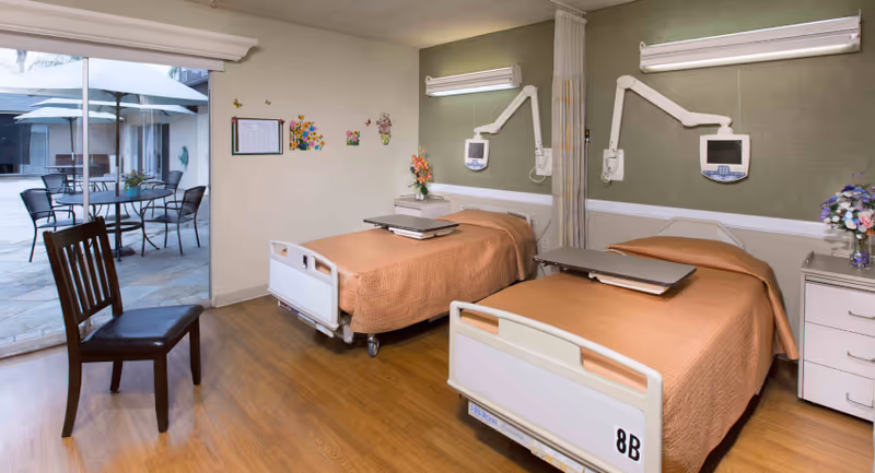 A shared patient room in a care center with two hospital beds covered in orange blankets. Each bed has an adjustable overbed table and a mounted medical monitor above the headboard. There is a wooden chair near a sliding glass door that opens to an outdoor patio with a table, chairs, and an umbrella. The room has wood flooring, light-colored walls with some decorations, and a nightstand with a flower vase.