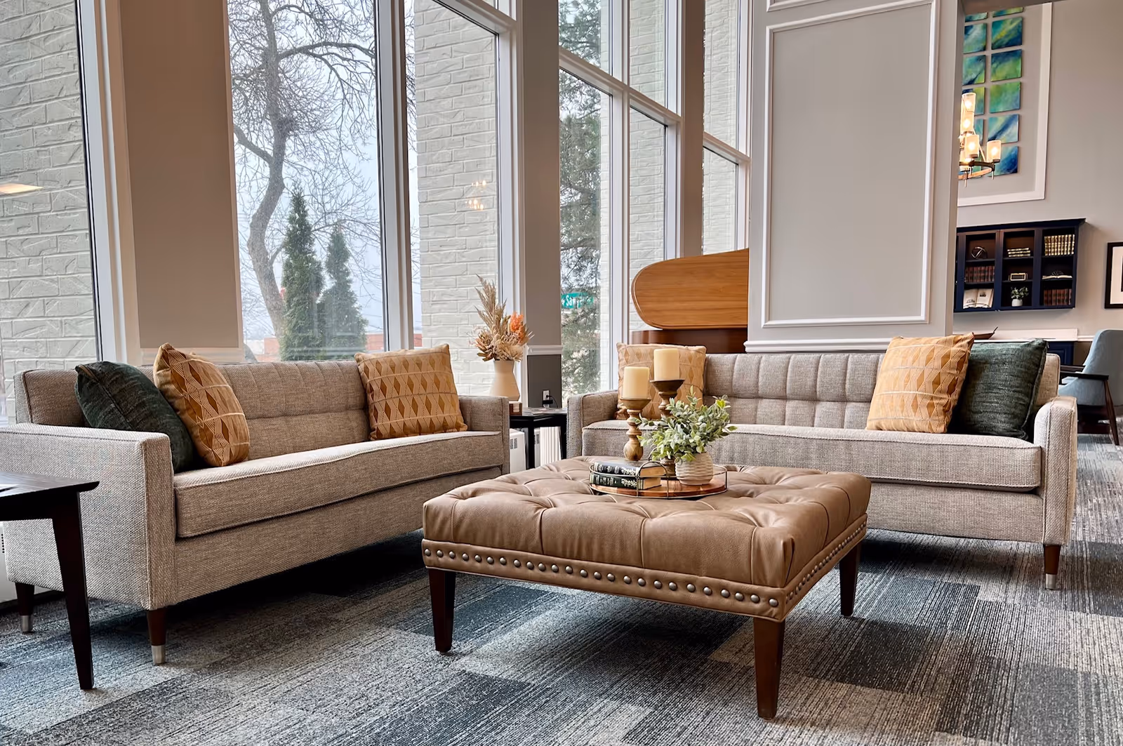 A bright and cozy living room area with two beige sofas adorned with patterned and solid throw pillows facing each other. Between them is a tufted leather ottoman with decorative candles, books, and a small plant on top. Large windows behind the sofas let in natural light and offer a view of trees outside. The room features a modern and inviting decor with a carpeted floor and a bookshelf in the background.
