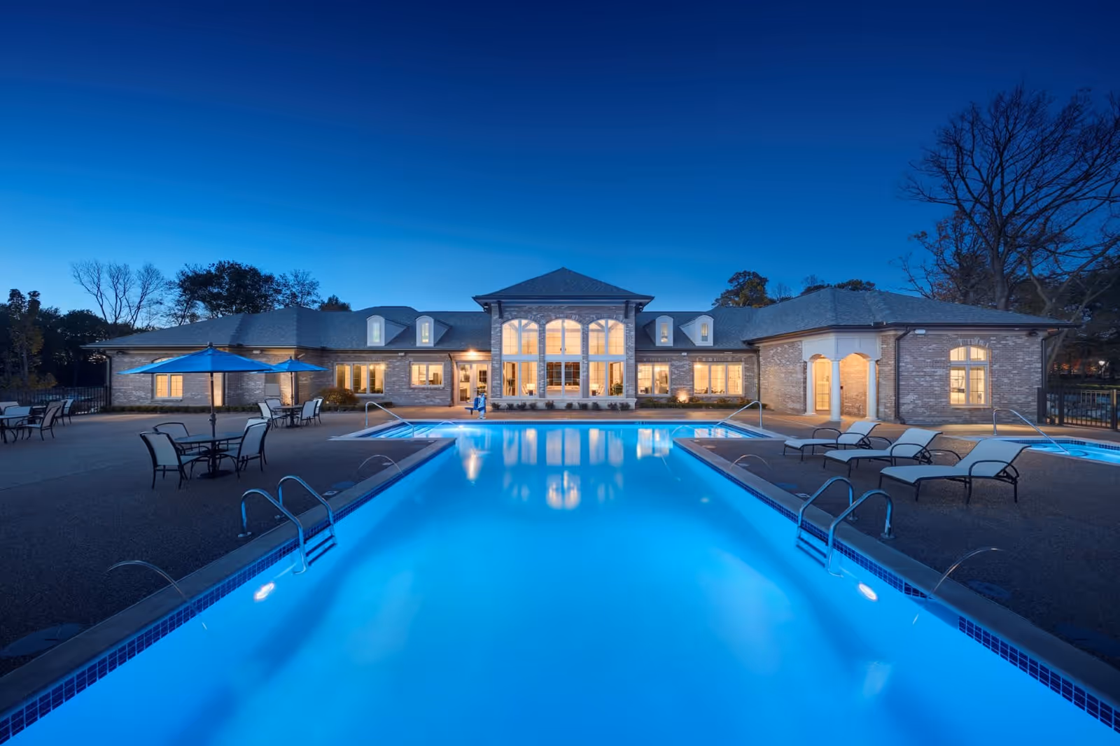 Evening view of a large outdoor swimming pool with clear blue water in front of a well-lit brick building with large windows. The pool area has lounge chairs and tables with umbrellas on the surrounding deck. Trees are visible in the background under a clear sky.