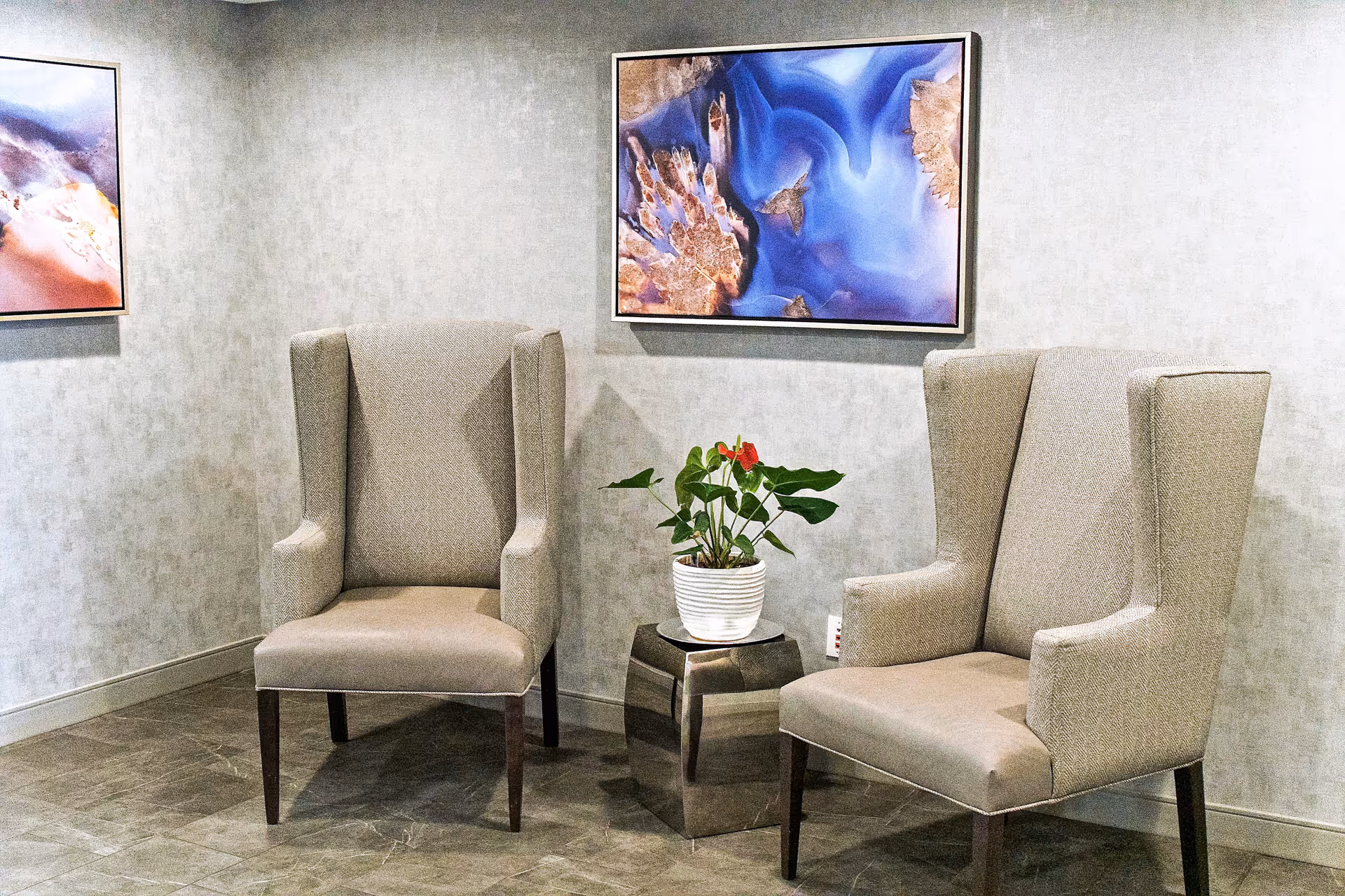 Two beige upholstered wingback chairs positioned on either side of a small metallic side table with a potted plant. The background features a light gray textured wall with two framed abstract artworks, one predominantly blue and the other with warm tones.