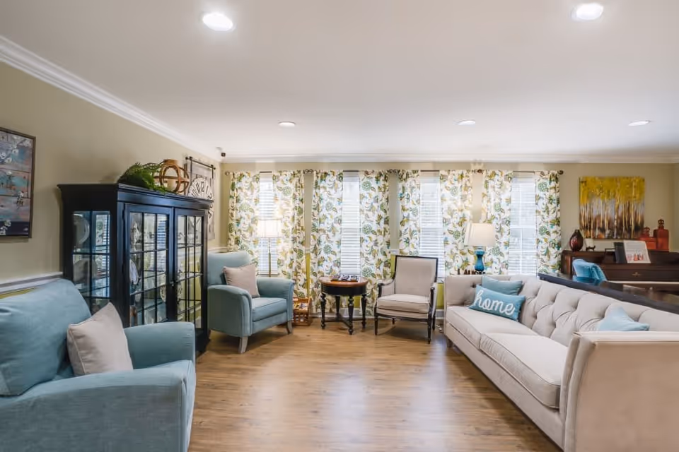 A cozy living room with light blue and beige upholstered chairs and a beige sofa with a blue pillow that says 'home'. The room has wooden flooring, floral patterned curtains on multiple windows, a black glass cabinet, a small round wooden table with a lamp, and a piano with decorative items on top.