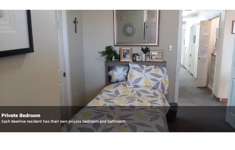 A small private bedroom with a single bed covered in a floral patterned bedspread with yellow and gray leaves. Above the bed is a mirror and a shelf holding framed photos and a small plant. The room has light-colored walls and a hallway visible to the right.