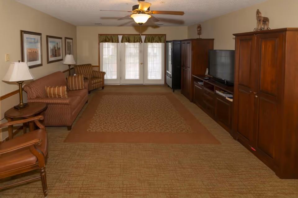 A cozy living room with a brown sofa, an armchair, a side table with a lamp, and a ceiling fan with light. There are three framed pictures on the wall above the sofa. Across from the seating area is a wooden entertainment center with a flat-screen TV and cabinets. French doors with sheer curtains and green valances let in natural light.