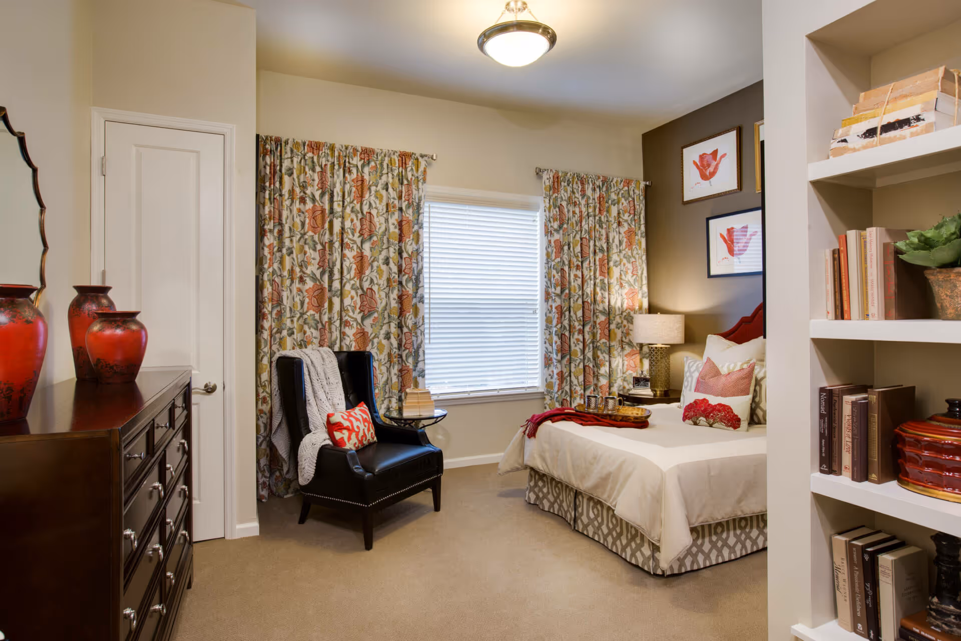 Well-decorated bedroom with a bed, armchair, dresser, bookshelf, and floral curtains.