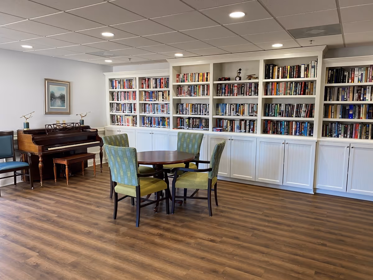 A cozy library room with a wooden floor, a round table surrounded by four green upholstered chairs, a wall of white bookshelves filled with books, and a wooden piano with a bench. A framed painting hangs on the wall above the piano.