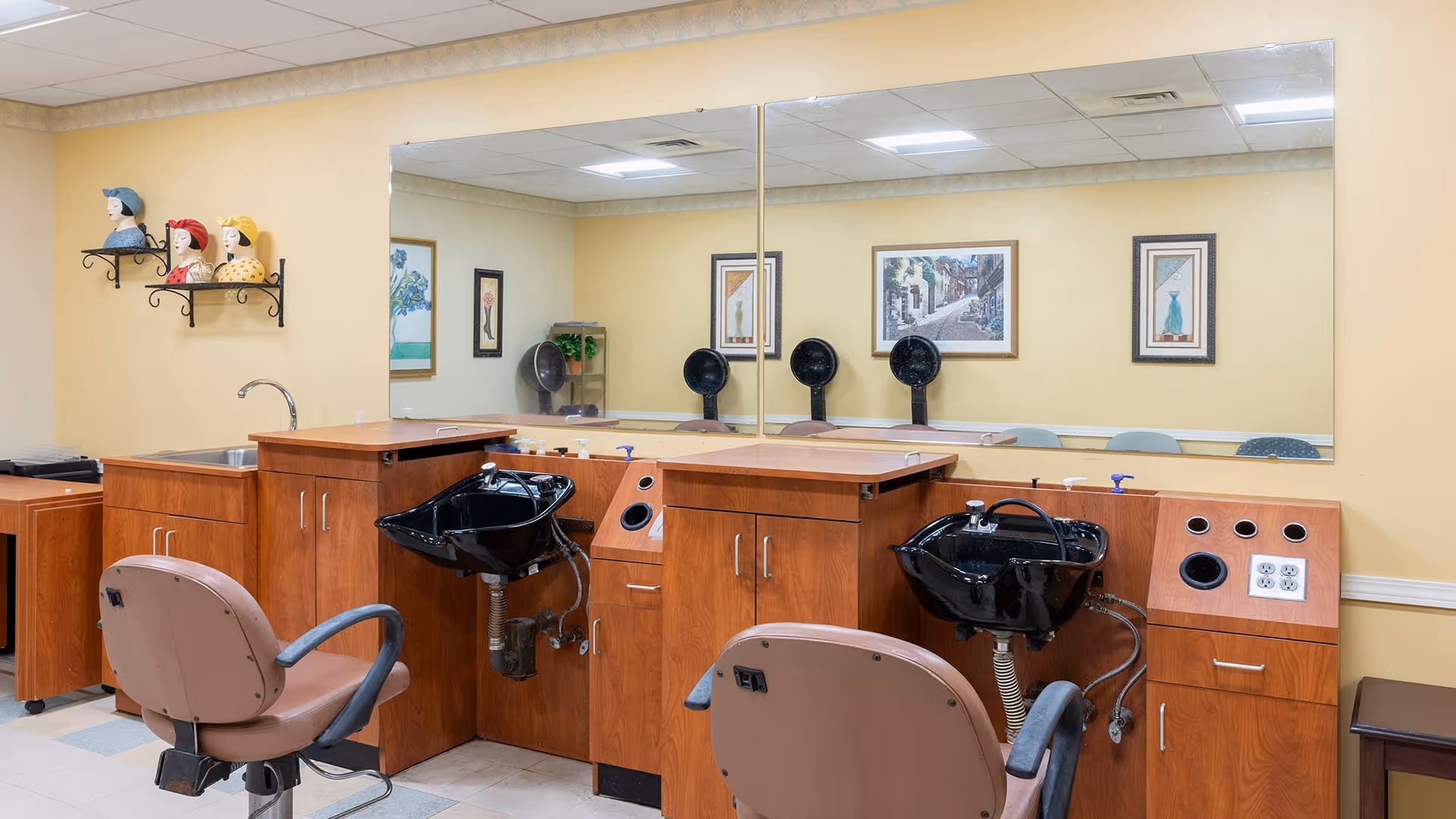 Interior view of a salon area in a senior living facility with two brown salon chairs facing black hair washing sinks mounted on wooden cabinetry. A large mirror covers the wall behind the sinks, reflecting framed artwork and additional salon equipment. The walls are painted light yellow, and decorative ceramic head sculptures are displayed on a wall-mounted shelf.