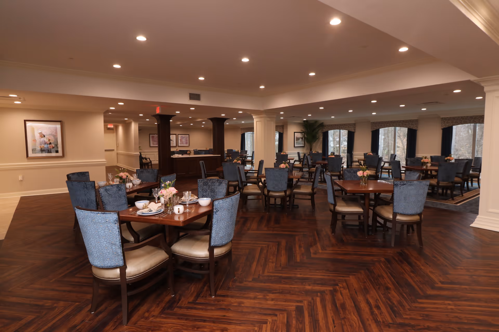A spacious dining room with multiple wooden tables and blue upholstered chairs arranged neatly. Each table has a small floral centerpiece and place settings. The room features dark wood flooring, cream-colored walls, recessed lighting, and large windows with dark curtains allowing natural light to enter.