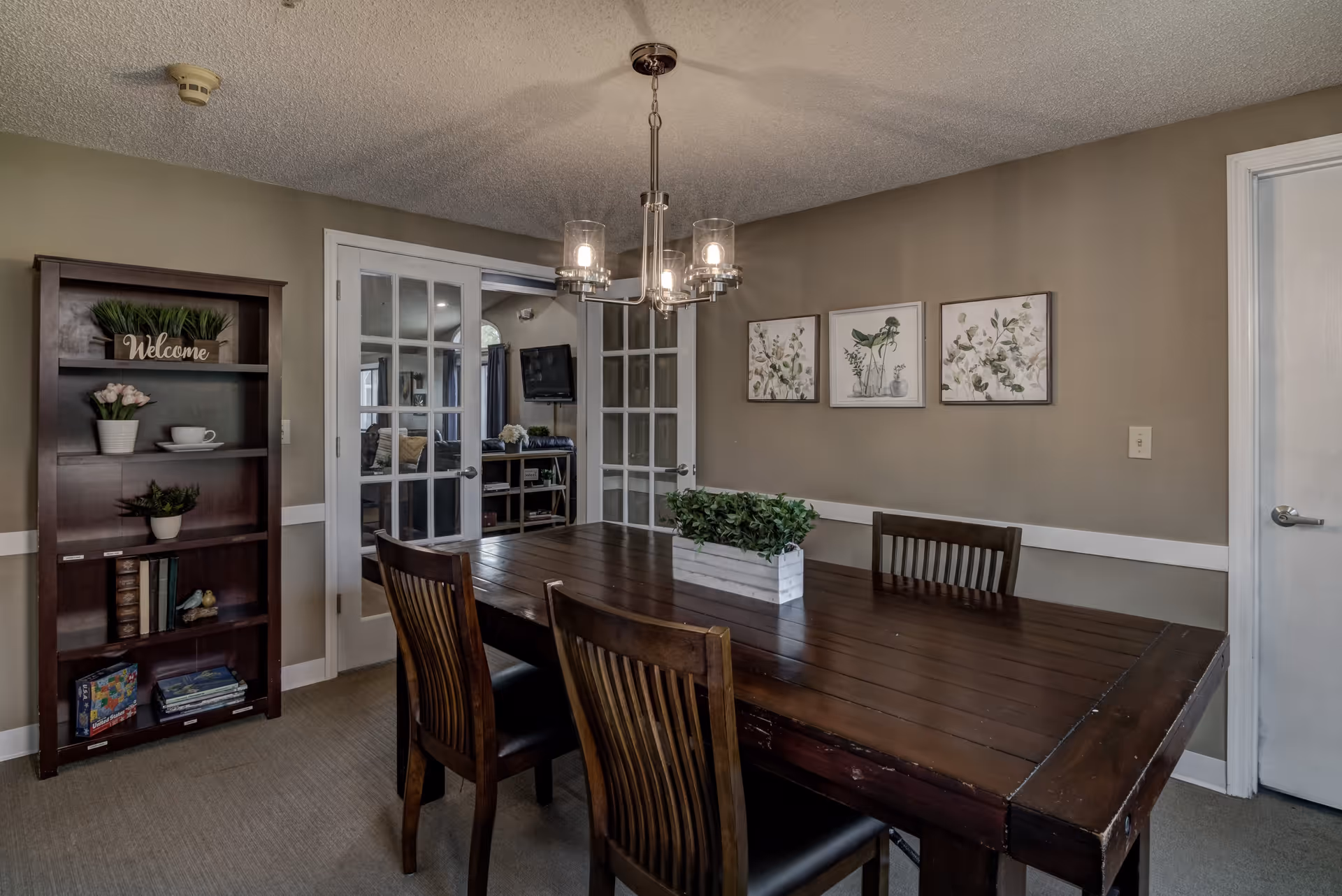 A dining room with a dark wooden table and four matching chairs. A rectangular planter with green plants is centered on the table. On the left side, there is a dark wooden bookshelf with decorative items including a 'Welcome' sign, plants, and books. The walls are painted beige and decorated with three framed botanical prints. French doors with glass panes lead to another room with a TV and additional seating. A modern chandelier with four lights hangs above the table.