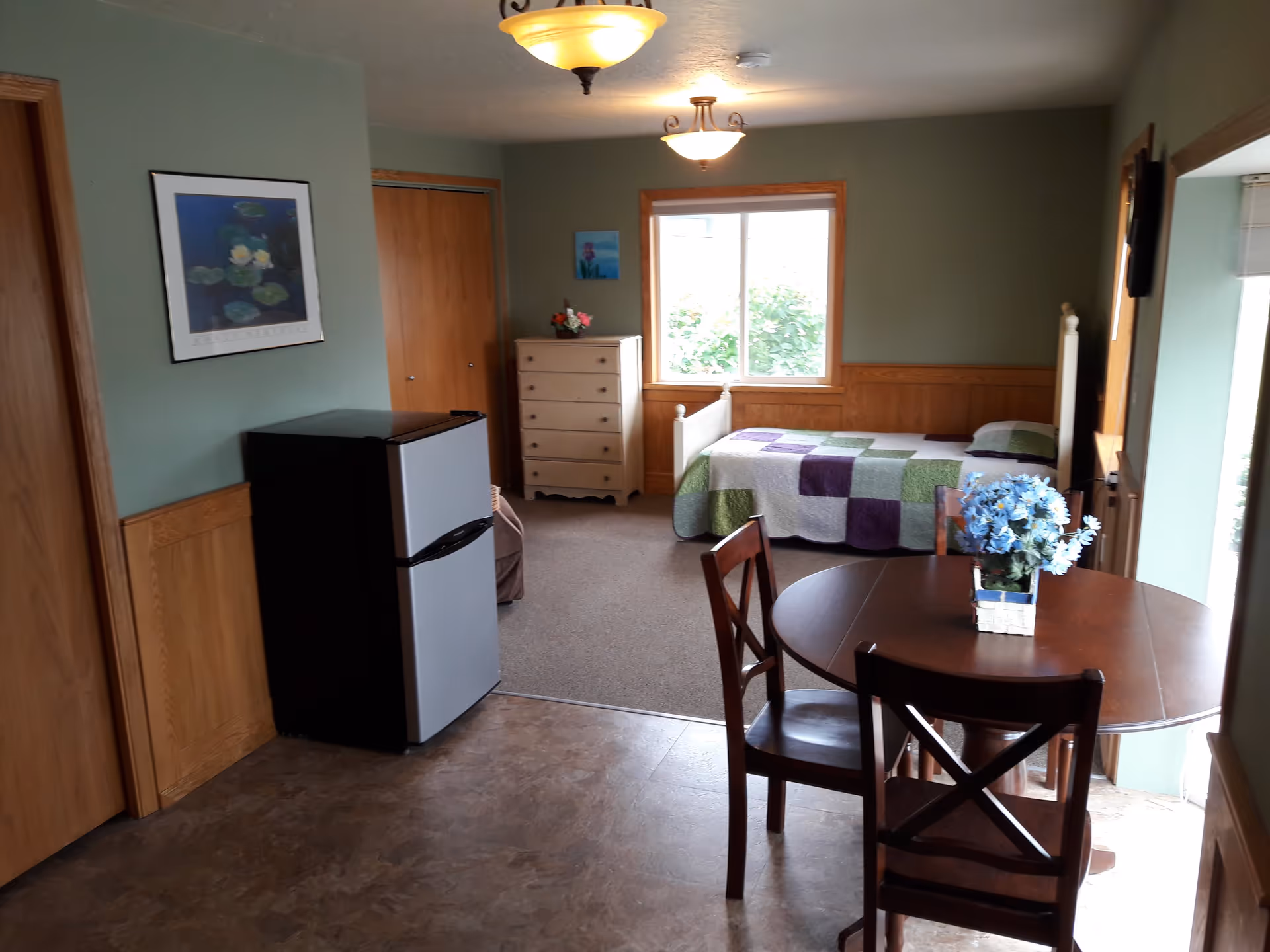Interior view of a room in Valley Pines Retirement Home featuring a small dining table with two chairs and a vase of blue flowers, a mini refrigerator, a dresser, a single bed with a patchwork quilt, and a window letting in natural light.