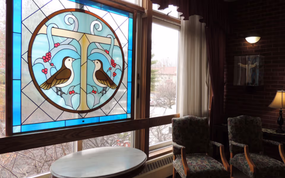 A cozy interior corner of a room with two patterned armchairs and a small round table in front of large windows. One window features a stained glass panel with a design of two birds perched on a stylized letter T with blue and red accents. The room has brick walls, a wall-mounted light fixture, a table lamp, and curtains partially drawn to the side.