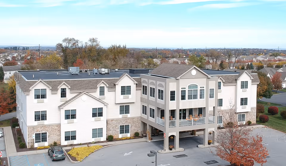 Three-story light-colored senior living building with a covered entrance/porte-cochere, balconies, parking lot, and autumn trees surrounding it.