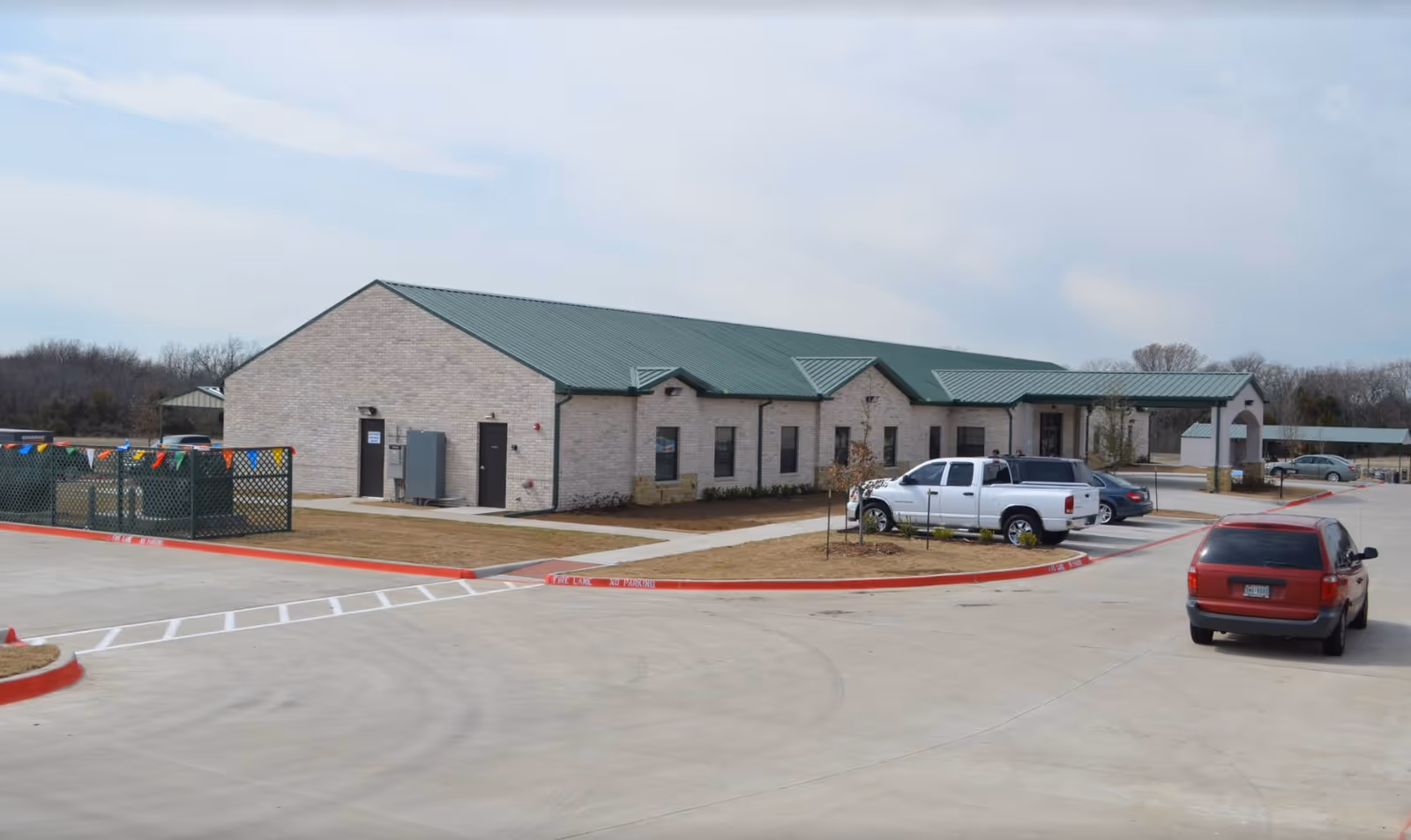Exterior view of a single-story brick building with a green metal roof, surrounded by a parking lot with several parked vehicles and a small fenced area with colorful flags. The sky is partly cloudy.