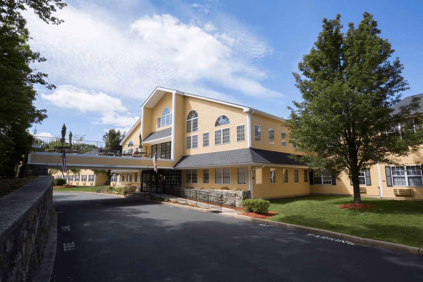 Exterior view of a large yellow senior living facility building with multiple windows, a covered entrance, and a connecting bridge. There are trees and a well-maintained lawn surrounding the building under a partly cloudy sky.
