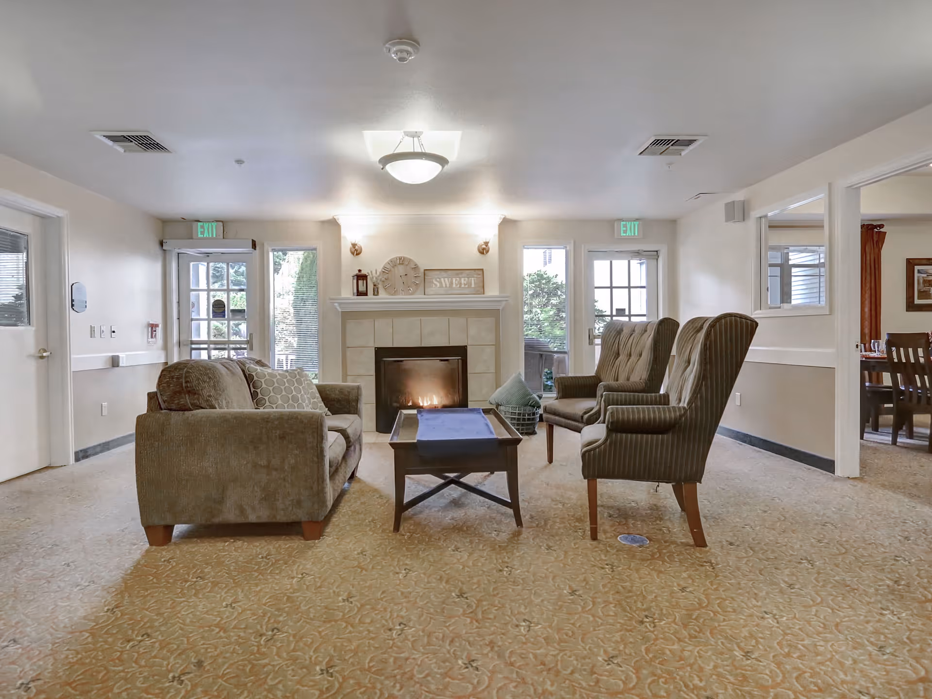 A cozy senior living facility common area with a fireplace in the center. There is a brown sofa on the left and two striped armchairs on the right, arranged around a wooden coffee table. The fireplace mantel has decorative items including a clock and a sign that says 'SWEET'. Large windows and glass doors allow natural light to enter the room.