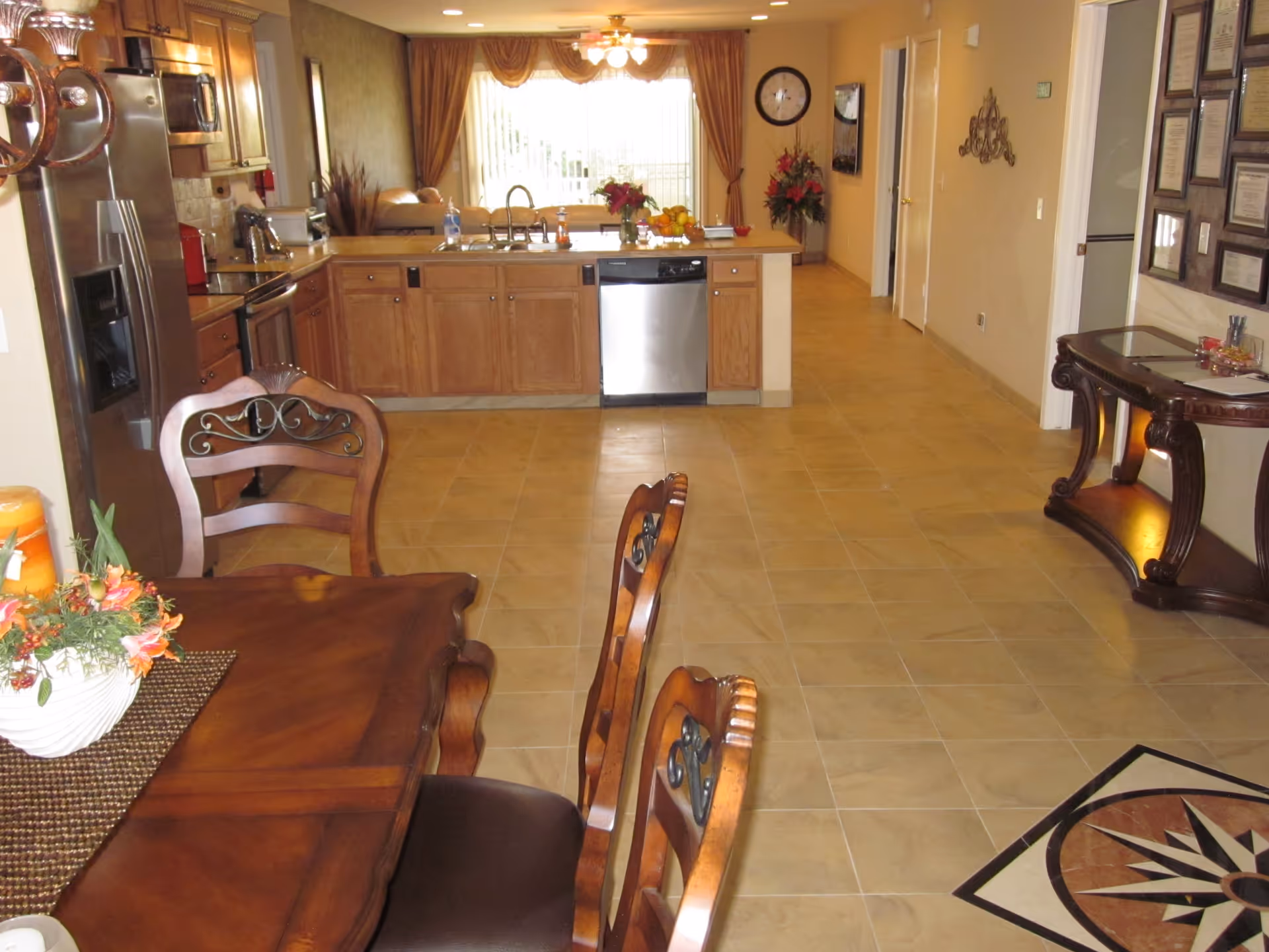 Interior view of a spacious kitchen and dining area in an assisted living facility. The kitchen features wooden cabinets, a stainless steel refrigerator, dishwasher, and a sink on a central island. The dining area has a wooden table with chairs and a decorative centerpiece. The floor is tiled, and there is a hallway leading to other rooms. The room is well-lit with natural light coming through large windows with curtains.