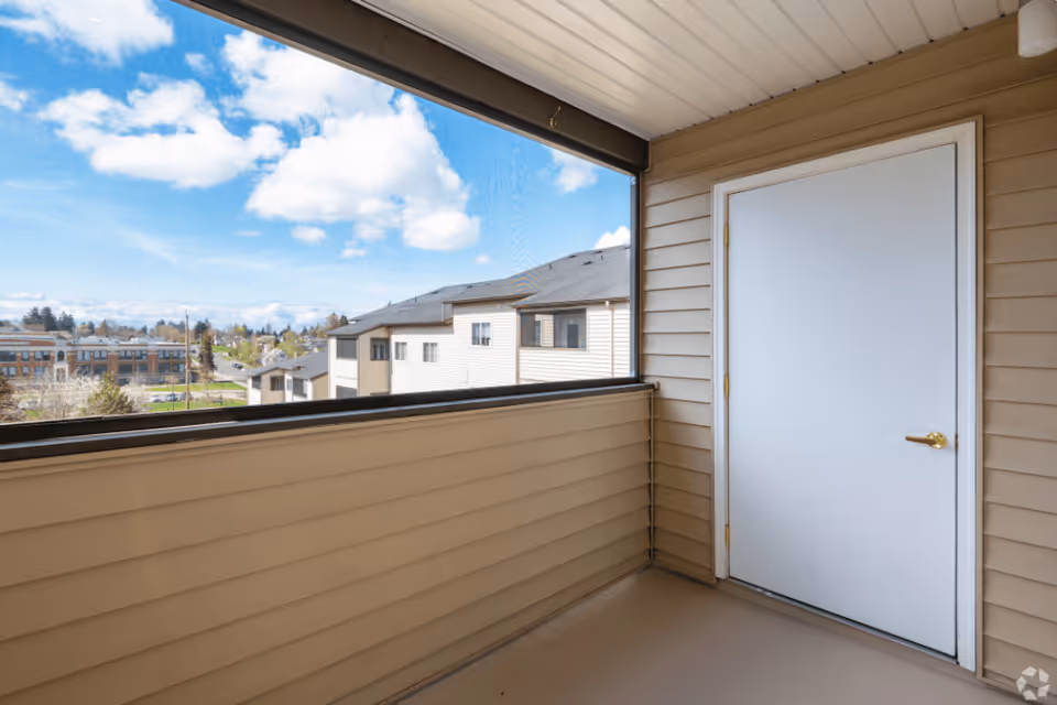 Covered apartment balcony with beige siding, a white door, and a view of neighboring buildings and a blue sky.