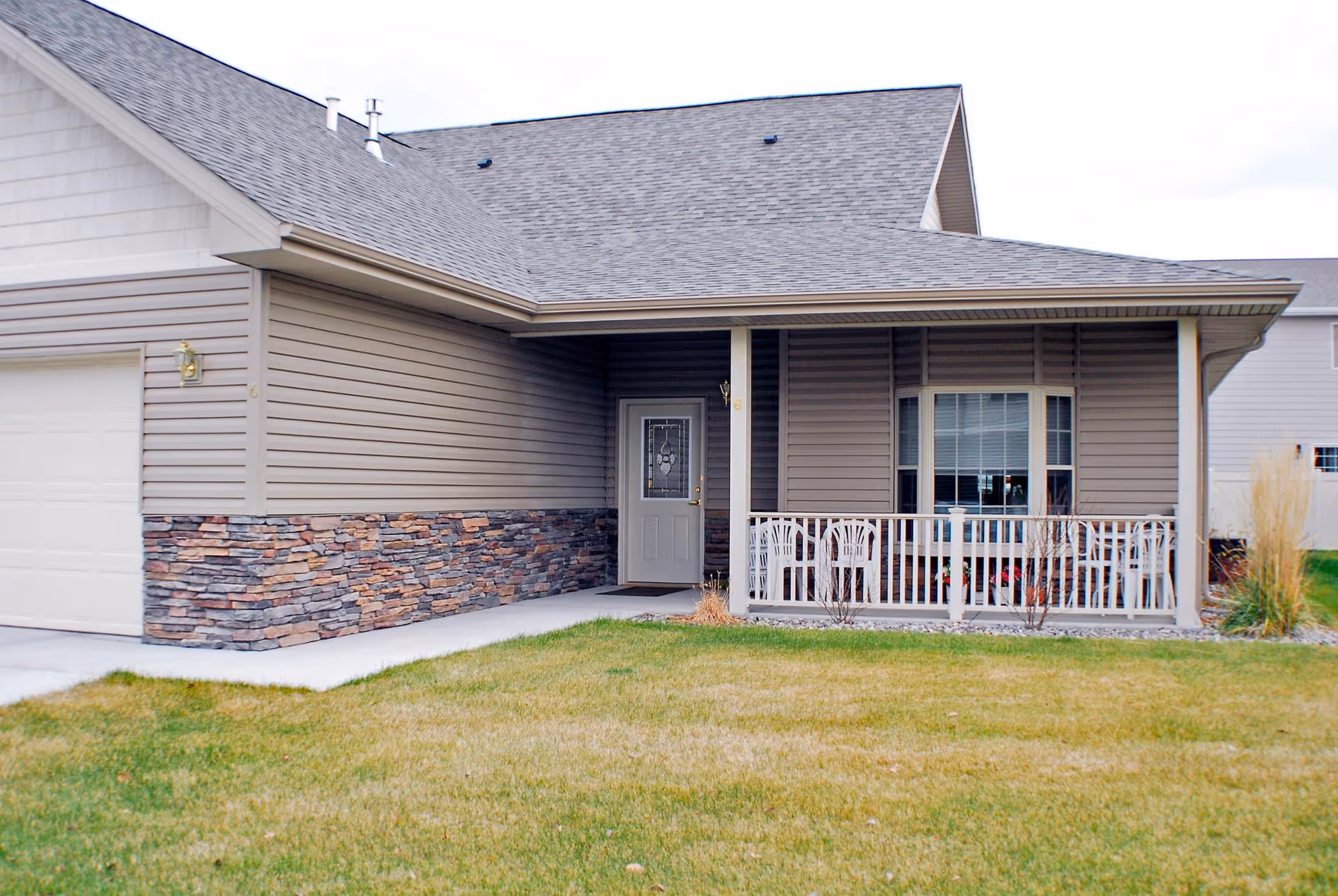 Exterior view of a single-story house with beige siding and stone accents. The house features a covered front porch with white railing and a glass front door. There is a well-maintained lawn in front of the house.