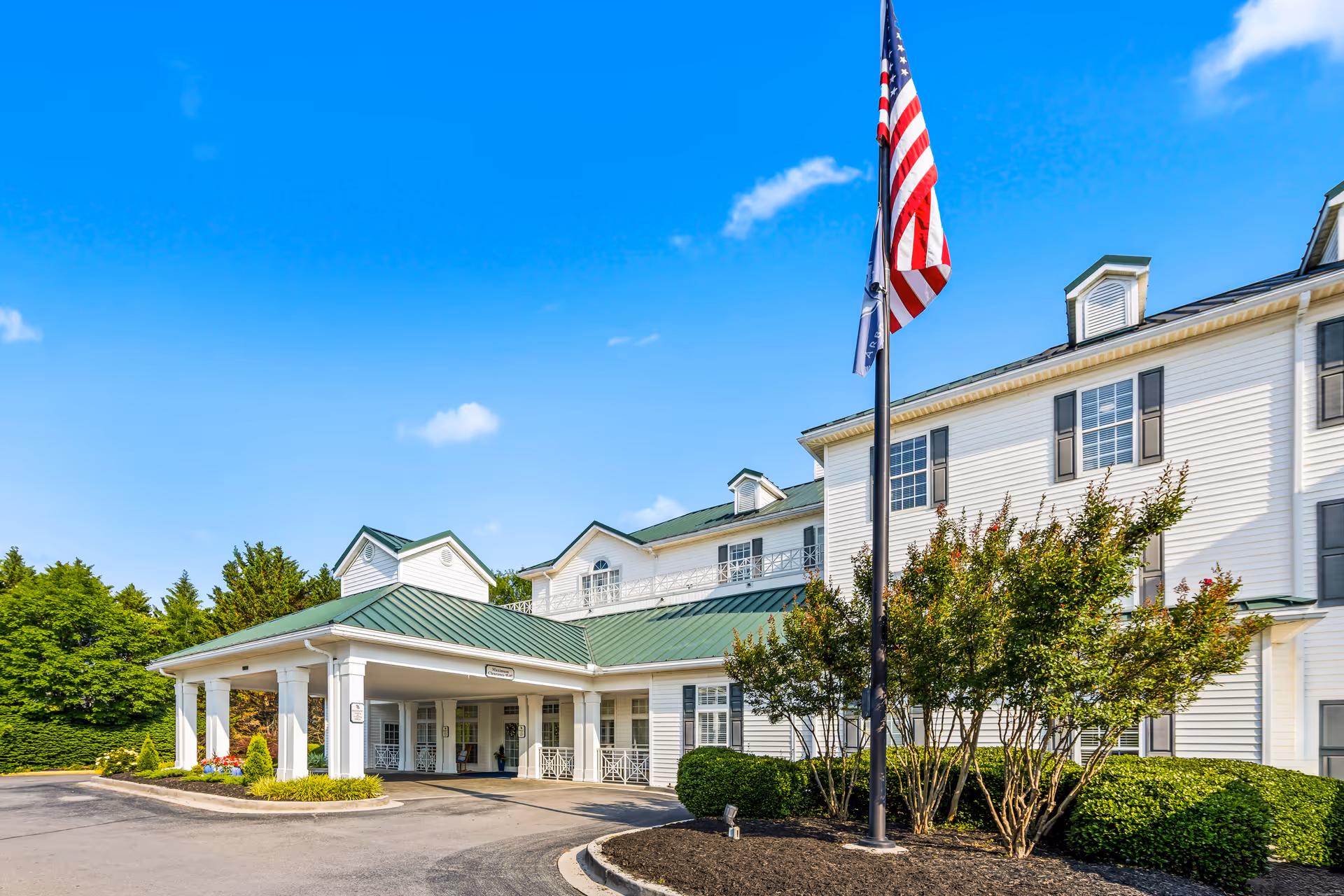 Front entrance of a white multi-story senior living building with a covered porte-cochere, landscaping, and an American flag.