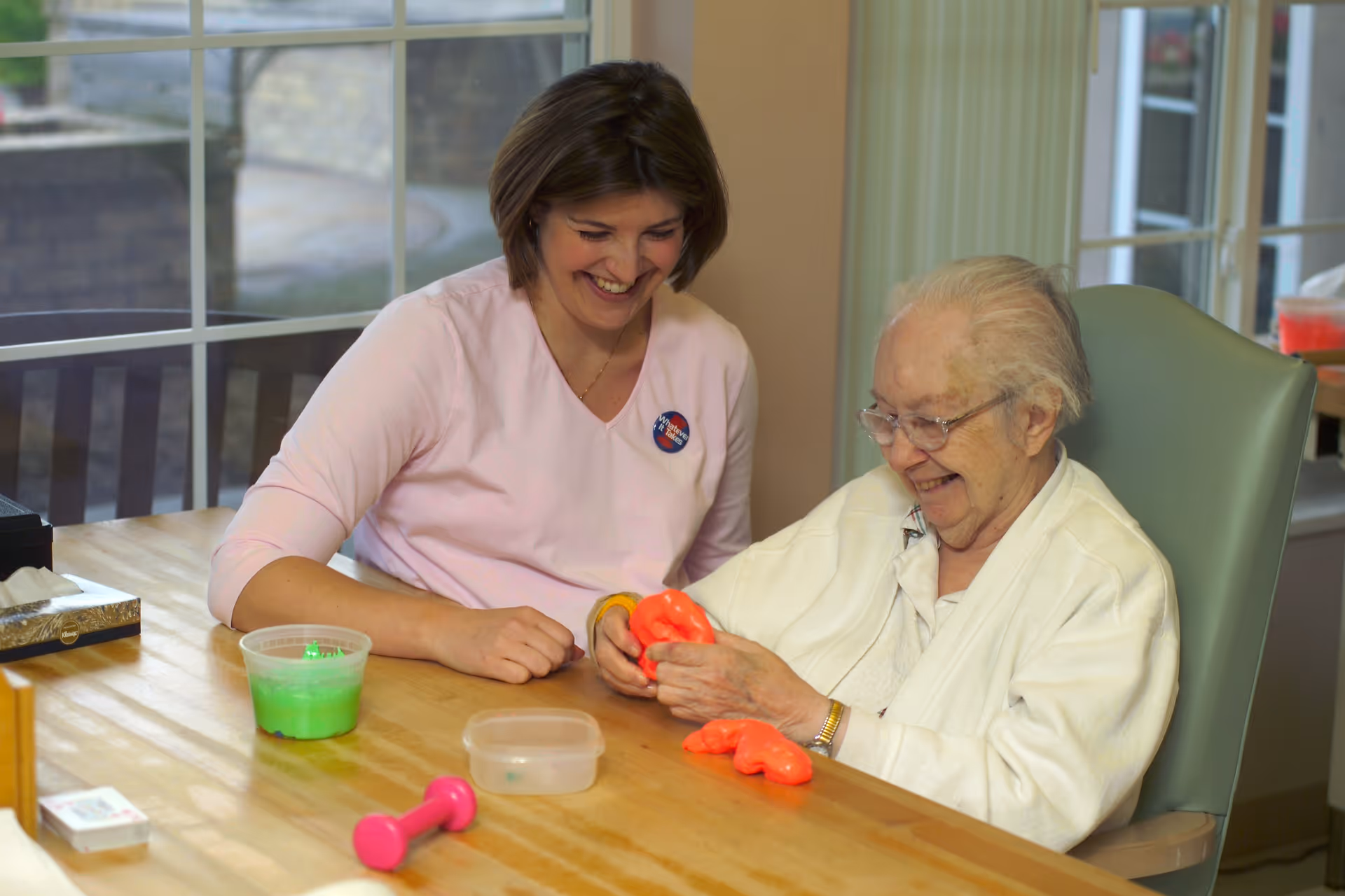 An elderly woman and a younger woman sitting at a wooden table, smiling and playing with bright orange therapy putty. The elderly woman is seated in a green chair, wearing glasses and a white cardigan, while the younger woman wears a light pink top and a badge. On the table are a pink dumbbell, a container with green putty, a small plastic box, and a tissue box. Large windows are in the background.
